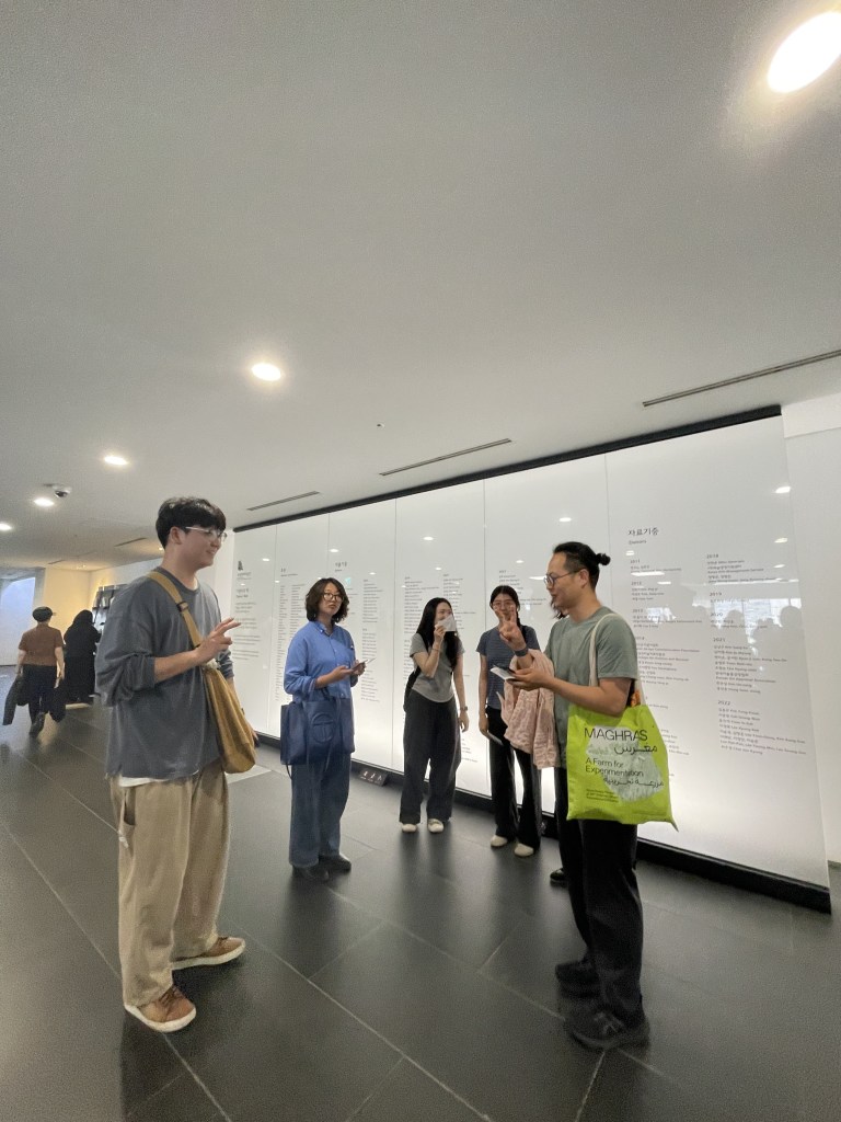 한국어 대체 텍스트: 여섯 명의 참여자가 국립현대미술관 로비의 아크릴 패널 앞에 반원 대형으로 서서 대화를 나누고 있습니다.
English Alt Text: A group of six people engaged in conversation standing in front of an information wall with text at the lobby of MMCA Seoul.