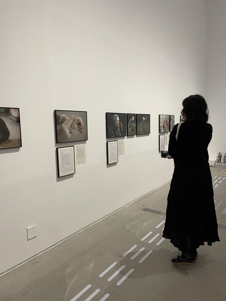 한국어 대체 텍스트: 검은 옷을 입은 여성 한 명이 전시실에서 작품을 감상하고 있습니다. 여성의 맞은 편에 있는 흰 벽에는 가로로 길쭉한 직사각형의 사진 작품이 검은색 액자에 담겨 눈높이보다 약간 아래에 걸려 있습니다. 어떤 사진 작품 밑에는 마찬가지로 검은색 액자에 담긴 손으로 쓴 편지가 함께 걸려 있습니다. 이 이미지에는 총 여덟 점의 사진과 세 점의 편지가 보입니다.
English Alt Text: A person dressed in black stands in front of a gallery wall, observing framed photographs and hand written letters in an art exhibition. There are eight photographs and three letters framed and hung on the wall.