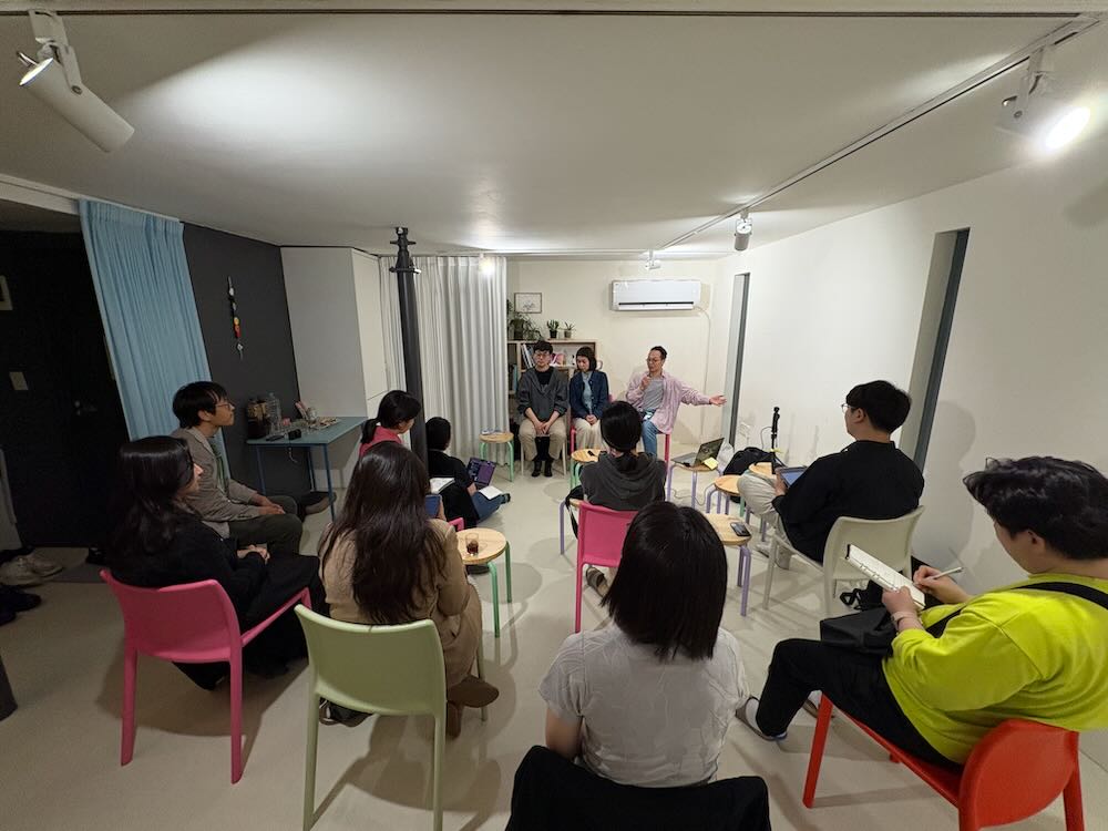 한국어 대체 텍스트: 실내 공간에서 진행되는 공개강연 모습. 흰색 벽과 회색 벽으로 구성된 현대적인 실내에 다양한 색깔의 플라스틱 의자들(분홍, 노랑, 주황, 빨강 등)이 원형으로 배치되어 있다. 약 15명의 참석자들이 의자에 앉아 강연을 듣고 있으며, 앞쪽에는 세 명의 강연자가 앉아 있다. 천장에는 트랙 조명이 설치되어 있고, 뒤쪽에는 책장과 식물들이 보인다.
English Alt Text: An open lecture taking place in an indoor space. A modern interior with white and gray walls features colorful plastic chairs (pink, yellow, orange, red, etc.) arranged in a circle. Approximately 15 participants are seated listening to the lecture, with three speakers seated at the front. Track lighting is installed on the ceiling, and bookshelves and plants are visible in the background.