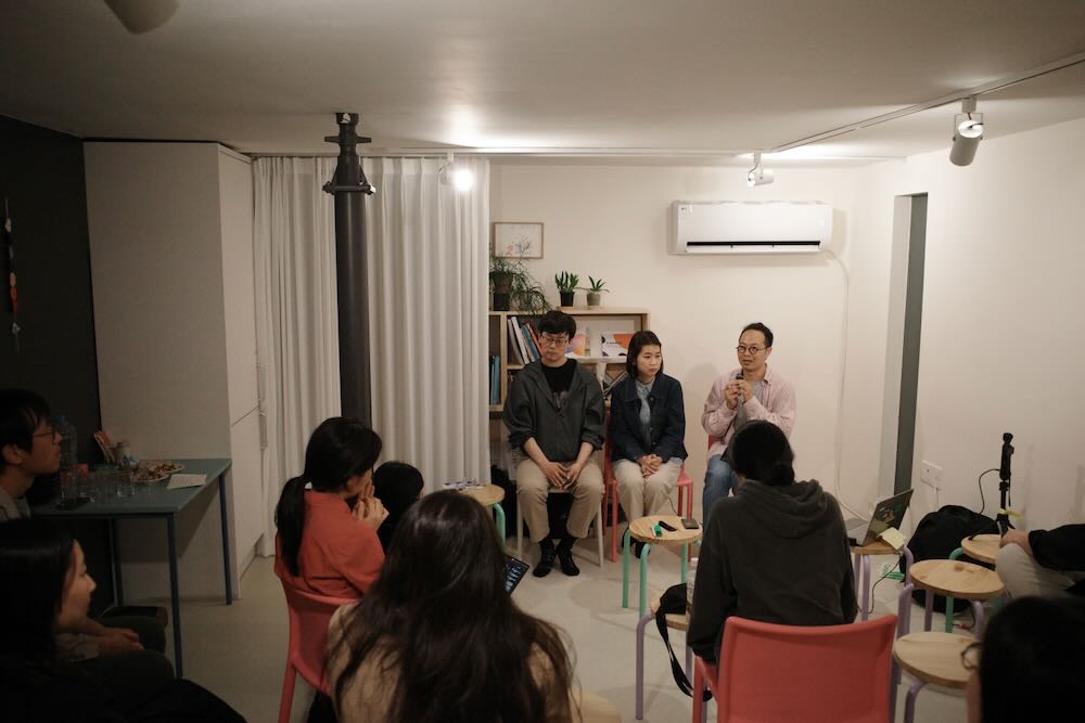 한국어 대체 텍스트:
같은 실내 공간에서의 강연 장면을 다른 각도에서 촬영한 모습. 참석자들이 다채로운 색상의 의자에 앉아 앞쪽을 바라보고 있으며, 세 명의 강연자 중 한 명이 마이크를 들고 발언하고 있다. 배경에는 책장, 에어컨, 흰색 커튼이 있는 창문이 보인다.
English Alt Text:
The same lecture scene captured from a different angle in the same indoor space. Participants are seated on colorful chairs facing forward, with one of the three speakers holding a microphone and speaking. The background shows bookshelves, an air conditioner, and a window with white curtains.