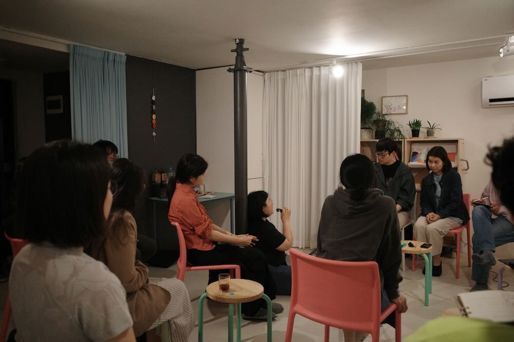 한국어 대체 텍스트:
강연 공간의 기술 장비 설치 모습. 흰색 벽 앞에 다채로운 색상의 플라스틱 스툴들이 쌓여 있고, 그 위에 노트북과 마이크가 설치되어 있다. 노트북 화면에는 화상회의 프로그램이 실행되어 있어 온라인 참여자들을 위한 스트리밍 준비가 되어 있음을 보여준다. 양쪽에는 참석자들의 다리가 일부 보이며, 바닥에는 가방과 자료들이 놓여 있다.
English Alt Text:
The technical equipment setup in the lecture space. Colorful plastic stools are stacked in front of a white wall, with a laptop and microphone installed on top. The laptop screen shows a video conferencing program running, indicating preparation for streaming for online participants. Parts of participants' legs are visible on both sides, and bags and materials are placed on the floor.