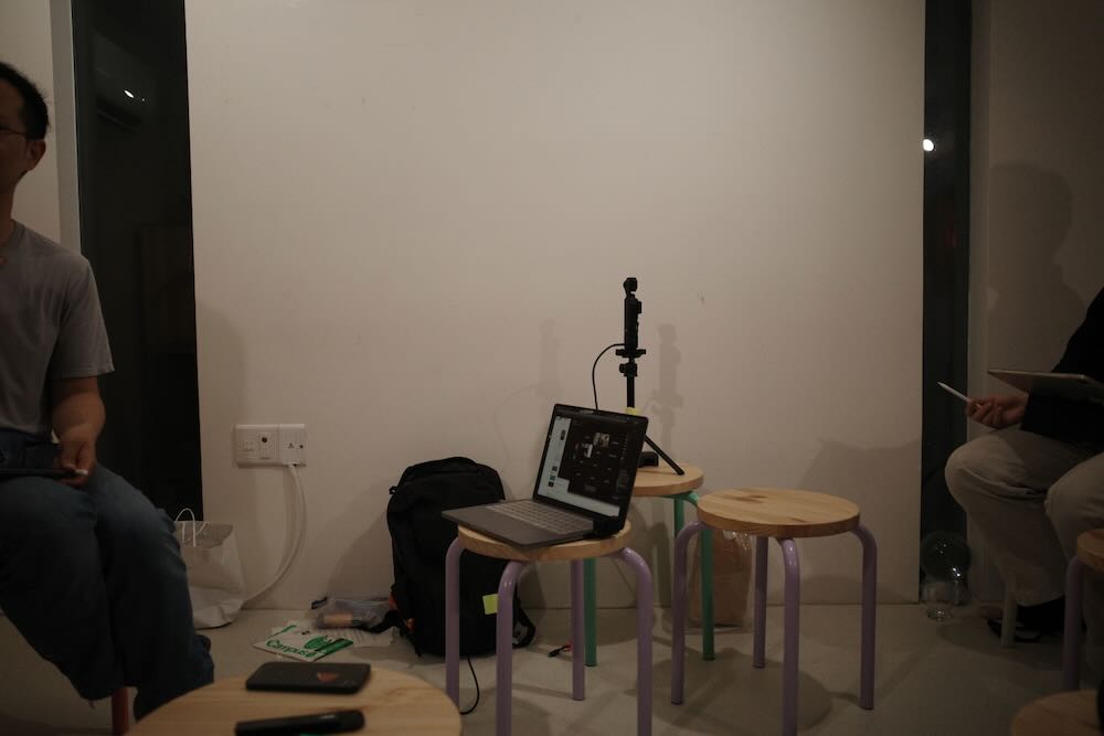 한국어 대체 텍스트:
강연이 진행 중인 실내 공간의 모습. 참석자들이 다양한 색깔의 플라스틱 의자에 앉아 대화를 나누고 있다. 왼쪽에는 청록색 커튼이 있는 창문과 검은색 벽이 보이고, 오른쪽에는 흰색 커튼, 책장, 그리고 식물들이 배치되어 있다. 중간에는 검은색 기둥이 있으며, 참석자들은 편안한 분위기에서 토론하고 있다. 한 사람이 메모를 하고 있고, 작은 원형 테이블 위에 물컵이 놓여 있다.
English Alt Text:
An indoor space where a lecture is in progress. Participants are seated on various colored plastic chairs, engaged in conversation. On the left, there's a window with turquoise curtains and a black wall, while on the right, white curtains, bookshelves, and plants are arranged. A black pillar stands in the middle, and participants are discussing in a relaxed atmosphere. One person is taking notes, and water cups are placed on a small round table.