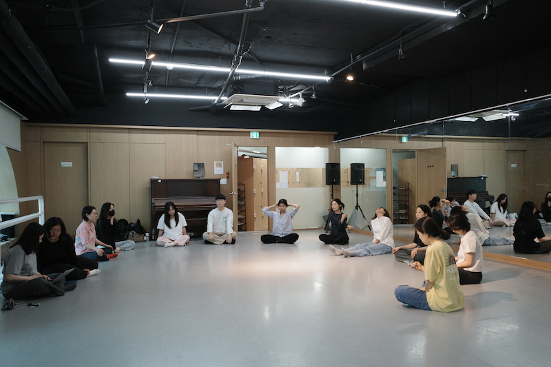 한국어 대체 텍스트: 연습실에서 참여자들이 원형으로 둘러앉아 워크숍에 참여하고 있는 모습. 편안한 복장의 참여자들이 바닥에 앉아 있으며, 일부는 스트레칭을 하거나 몸을 움직이고 있다.
English alternative text: Participants sitting in a circle in the practice room during the workshop. Dressed in comfortable clothes, participants are seated on the floor, with some stretching or moving their bodies.