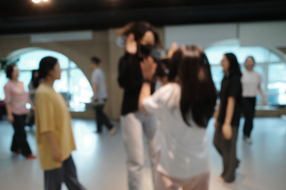 한국어 대체 텍스트: 연습실에서 여러 참여자들이 움직임 워크숍에 참여하고 있는 모습. 사진이 약간 흐릿하게 처리되어 움직임의 역동성이 강조되고 있으며, 참여자들이 팔을 들거나 다양한 동작을 취하고 있다.
English alternative text: Multiple participants engaged in a movement workshop in the practice room. The photo is slightly blurred, emphasizing the dynamism of movement, with participants raising their arms and making various gestures.