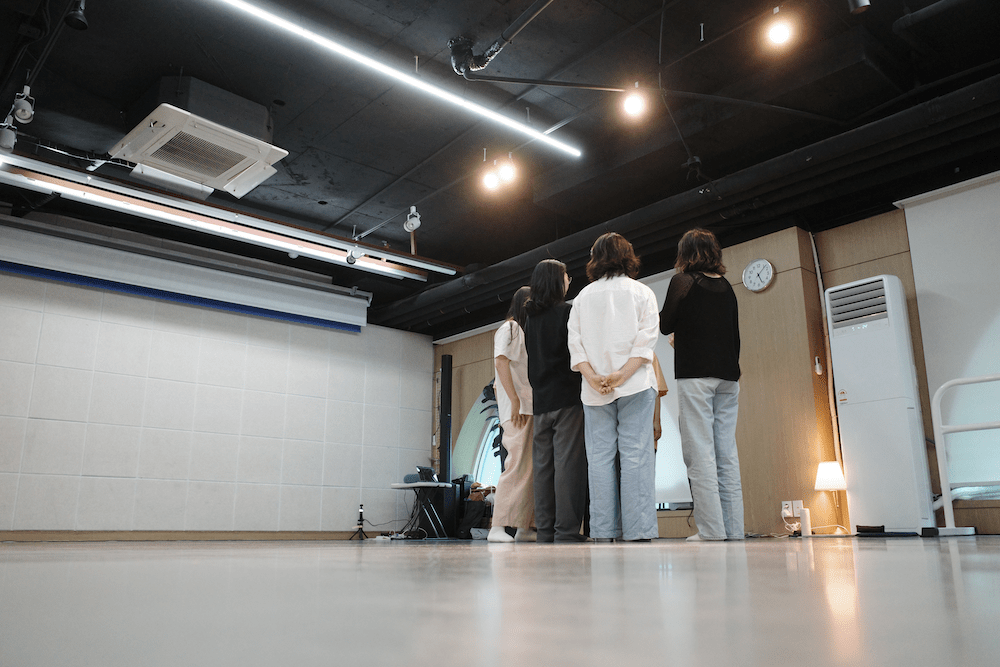 한국어 대체 텍스트: 네 명의 참여자가 연습실에서 나란히 서서 같은 방향을 바라보고 있는 모습. 뒷모습이 보이며, 편안한 복장을 입고 차분한 자세로 서 있다.
English alternative text: Four participants standing side by side in the practice room, facing the same direction. Viewed from behind, they are wearing comfortable clothes and standing in calm postures.