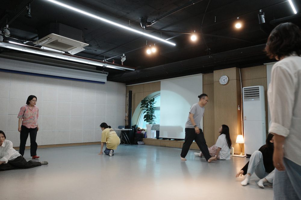 한국어 대체 텍스트: 참여자들이 연습실 바닥에서 다양한 높이의 움직임을 탐구하고 있는 모습. 일부는 서 있고, 일부는 무릎을 꿇거나 바닥에 앉아 있으며, 한 명은 걷고 있다. 공간 전체에서 각자 다른 수준의 움직임이 동시에 일어나고 있다.
English alternative text: Participants exploring movements at various heights in the practice room. Some are standing, others are kneeling or sitting on the floor, and one person is walking. Different levels of movement are happening simultaneously throughout the space.